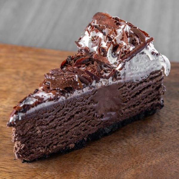 A slice of rich chocolate cheesecake with whipped cream and chocolate shavings on a wooden board.